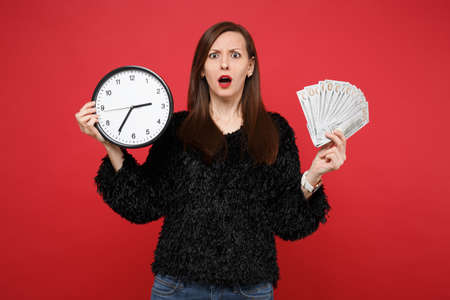 Perplexed young woman in black fur sweater holding round clock, fan of money in dollar banknotes cash money isolated on red background. People sincere emotions, lifestyle concept. Time is running outの写真素材