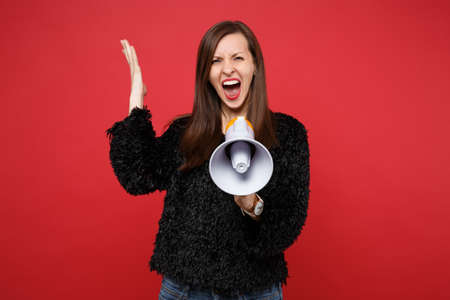 Portrait of irritated young woman in black fur sweater spreading hand, screaming on megaphone...