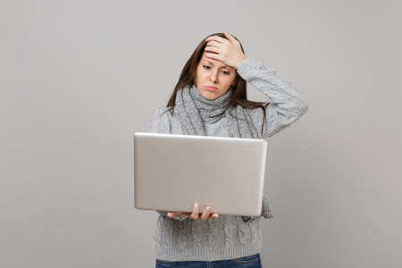 Upset young woman in gray sweater, scarf putting hand on forehead, working on laptop pc computer isolated on grey wall background. Healthy lifestyle, online treatment consulting, cold season conceptの写真素材