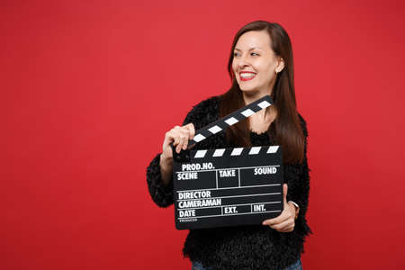 Smiling young woman in black fur sweater looking aside, holding classic black film making clapperboard isolated on red wall background. People sincere emotions, lifestyle concept. Mock up copy spaceの写真素材