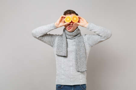 Crazy young man in gray sweater, scarf covering eyes with oranges fooling around isolated on grey background. Healthy fashion lifestyle people sincere emotions cold season concept. Mock up copy spaceの写真素材