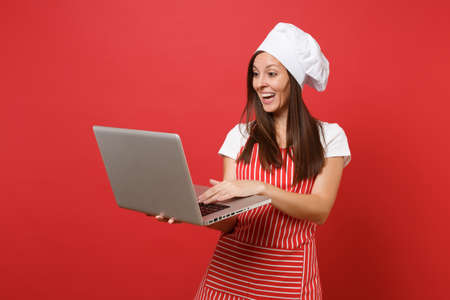 Housewife female chef cook or baker in striped apron, white t-shirt, toque chefs hat isolated on red wall background. Fun housekeeper woman looking for recipe in laptop pc. Mock up copy space conceptの写真素材