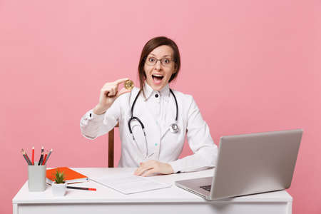Female doctor sits at desk works on computer with medical document hold coin in hospital isolated on pastel pink background. Woman in medical gown glasses stethoscope. Healthcare medicine conceptの写真素材