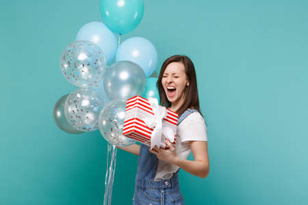 Crazy screaming girl hold red striped present box with gift ribbon celebrating with colorful air balloons isolated on blue turquoise background. Valentine's Day Women's Day, birthday, holiday conceptの写真素材