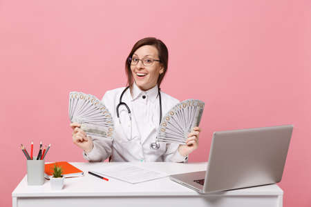 Female doctor sit at desk work on computer with medical document cash money in hospital isolated on pastel pink wall background. Woman in medical gown glasses stethoscope. Healthcare medicine conceptの写真素材