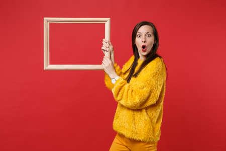 Portrait of shocked young woman in yellow fur sweater keeping mouth wide open, hold picture frame isolated on red background in studio. People sincere emotions, lifestyle concept. Mock up copy spaceの写真素材