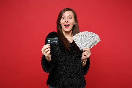 Surprised young woman in black fur sweater hold credit bank card fan of money in dollar banknotes cash money isolated on red background. People sincere emotions, lifestyle concept. Mock up copy spaceの写真素材