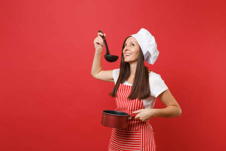 Housewife female chef cook or baker in striped apron white t-shirt toque chefs hat isolated on red wall background. Woman hold tasting empty stewpan black soup ladle dipper Mock up copy space conceptの写真素材