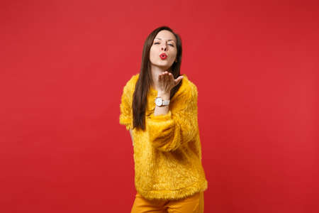 Portrait of tender pretty young woman in yellow fur sweater blowing, sending air kiss isolated on bright red wall background in studio. People sincere emotions, lifestyle concept. Mock up copy spaceの写真素材