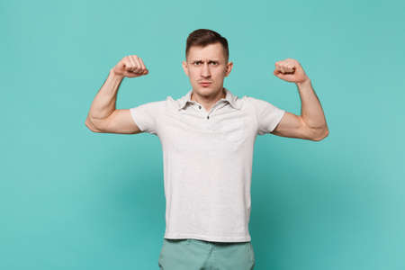 Portrait of powerful young man in casual clothes standing, showing biceps, muscles isolated on blue turquoise wall background in studio. People sincere emotions, lifestyle concept. Mock up copy spaceの写真素材