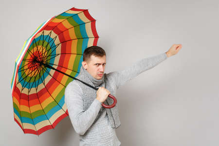 Upset young man in gray sweater, scarf holding colorful umbrella pointing hand isolated on grey background. Healthy fashion lifestyle, people sincere emotions, cold season concept. Mock up copy spaceの写真素材