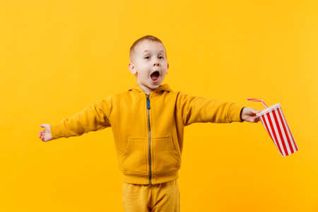 Little fun cheerful kid boy 3-4 years old wearing yellow clothes hold cup of soda isolated on orange wall background, children studio portrait. People, childhood lifestyle concept. Mock up copy spaceの写真素材