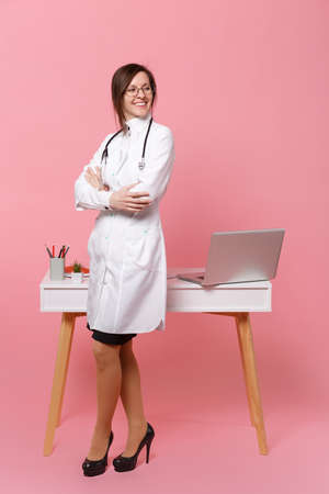 Cute female doctor stand in front of desk with pc computer, medical document in hospital isolated on pastel pink background. Woman in medical gown glasses stethoscope. Healthcare medicine conceptの写真素材