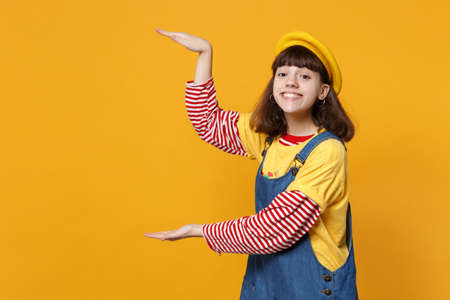 Portrait of smiling girl teenager in french beret and denim sundress pointing hands aside isolated...