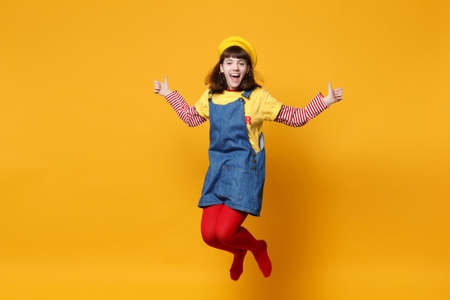 Laughing girl teenager in french beret, denim sundress showing thumbs up jumping and fooling around...