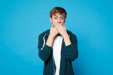 Portrait of shocked young man in casual clothes covering mouth with hands isolated on blue wall...