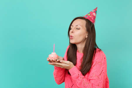 Stunning young girl in knitted pink sweater birthday hat with closed eyes blowing out candle on cake in hands isolated on blue background studio portrait. People lifestyle concept. Mock up copy spaceの写真素材