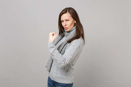 Funny young woman in gray sweater, scarf showing biceps, muscles isolated on grey wall background in...