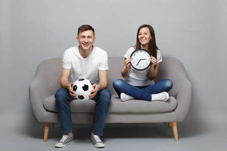 Fun couple woman man football fans in white t-shirt cheer up support favorite team with soccer ball holding round clock isolated on grey wall background. People sport family leisure lifestyle conceptの写真素材