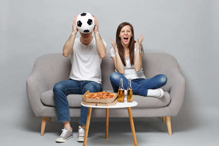 Screaming couple woman man football fans in white t-shirt cheer up support favorite team, covering face with soccer ball isolated on grey background. People emotions, sport family lifestyle conceptの写真素材