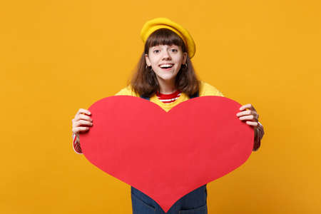 Excited girl teenager in french beret, denim sundress holding big empty blank red heart isolated on yellow wall background in studio. People sincere emotions, lifestyle concept. Mock up copy spaceの写真素材