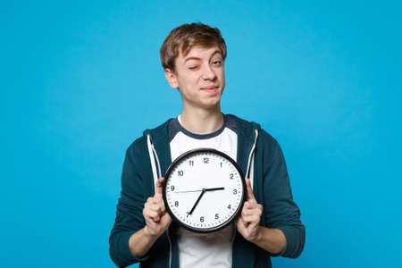 Portrait of funny young man in casual clothes blinking, holding round clock isolated on blue background in studio. Time is running out. People sincere emotions, lifestyle concept. Mock up copy spaceの写真素材