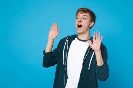 Portrait of surprised young man in casual clothes looking aside, rising hands, keeping mouth open isolated on blue background in studio. People sincere emotions, lifestyle concept. Mock up copy spaceの写真素材