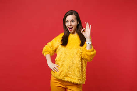 Amazed young woman in yellow fur sweater keeping mouth wide open, showing OK gesture isolated on bright red wall background in studio. People sincere emotions, lifestyle concept. Mock up copy spaceの写真素材