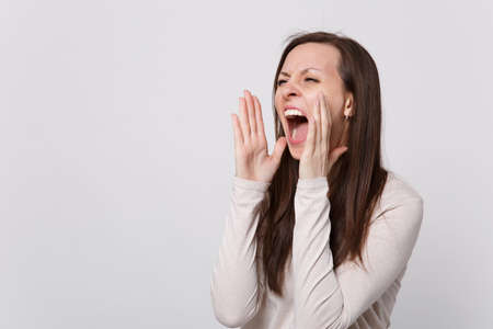 Irritated young woman in light clothes looking aside screaming with hand gesture near mouth isolated on white wall background in studio. People sincere emotions, lifestyle concept. Mock up copy spaceの写真素材