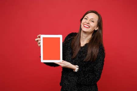 Happy young woman in black fur sweater holding tablet pc computer with blank empty screen isolated on red wall background in studio. People sincere emotions, lifestyle concept. Mock up copy spaceの写真素材