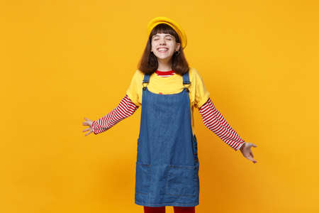 Portrait of funny girl teenager in french beret, denim sundress keeping eyes closed, spreading hands isolated on yellow wall background. People sincere emotions, lifestyle concept. Mock up copy spaceの写真素材
