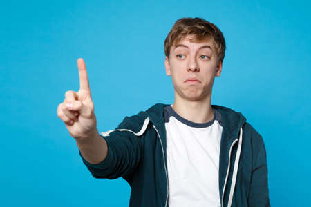 Young man in casual clothes touch something like push click on button, pointing at floating virtual screen isolated on blue background. People sincere emotions, lifestyle concept. Mock up copy spaceの写真素材