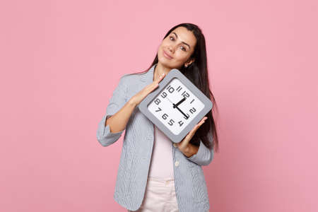 Portrait of smiling pretty young woman in striped jacket holding in hands square clock isolated on pink pastel wall background in studio. People sincere emotions lifestyle concept. Mock up copy spaceの写真素材