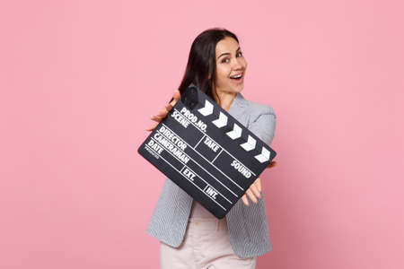 Cheerful young woman in striped jacket holding classic black film making clapperboard isolated on pink pastel wall background in studio. People sincere emotions, lifestyle concept. Mock up copy spaceの写真素材