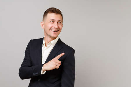 Smiling young business man in classic black suit, shirt looking, pointing index finger aside isolated on grey wall background in studio. Achievement career wealth business concept. Mock up copy spaceの写真素材