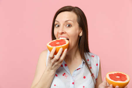 Excited funny young woman in summer clothes holding in hand, biting half of fresh ripe grapefruit...