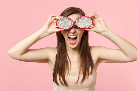 Screaming young woman covering eyes with halfs of fresh ripe pitahaya dragon fruit isolated on pink pastel wall background in studio. People vivid lifestyle relax vacation concept. Mock up copy spaceの写真素材