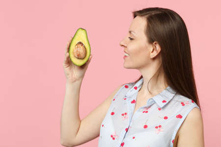 Side view of young woman in summer clothes holding fresh ripe green avocado fruit isolated on pink...