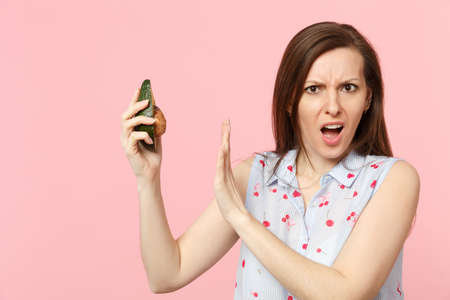 Disgusted young woman in summer clothes showing stop gesture with palm to green avocado fruit...