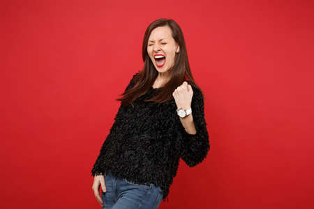 Overjoyed young woman in black fur sweater doing winner gesture, keeping eyes closed, screaming...