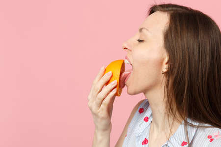 Side view of young woman in summer clothes keeping eyes closed licking half of fresh orange fruit isolated on pink pastel background. People vivid lifestyle relax vacation concept. Mock up copy spaceの写真素材
