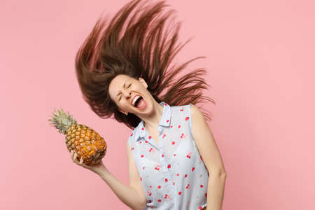 Crazy screaming young woman with flowing hair keeping eyes closed hold fresh ripe pineapple fruit isolated on pink pastel background. People vivid lifestyle relax vacation concept. Mock up copy spaceの写真素材