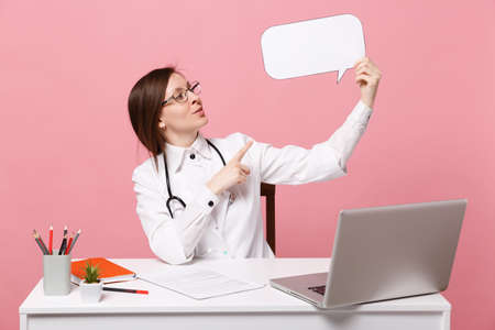 Female doctor sit at desk work on computer with medical document hold cloud in hospital isolated on pastel pink wall background. Woman in medical gown glasses stethoscope. Healthcare medicine conceptの写真素材