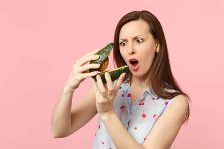 Shocked woman in summer clothes keeping mouth open holding two halfs of fresh ripe avocado fruit...