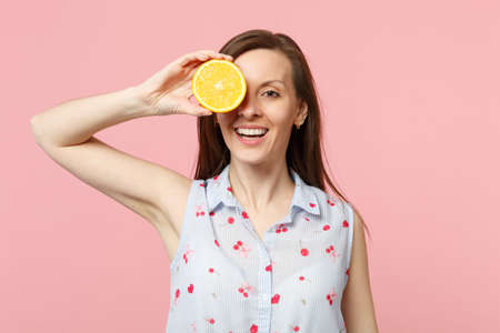 Smiling young woman in summer clothes holding covering eye with halfs of fresh ripe orange fruit...