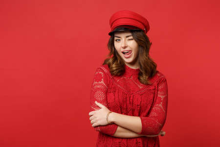 Portrait of blinking sexy young woman in lace dress, cap looking camera showing tongue isolated on bright red wall background in studio. People sincere emotions lifestyle concept. Mock up copy spaceの写真素材