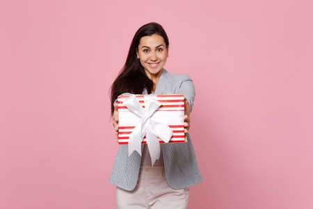 Joyful young woman holding red striped present box with gift ribbon isolated on pink pastel...