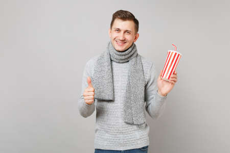 Handsome young man in gray sweater, scarf holding plastic cup of cola or soda, showing thumb up isolated on grey background. Healthy fashion lifestyle, people sincere emotions, cold season conceptの写真素材