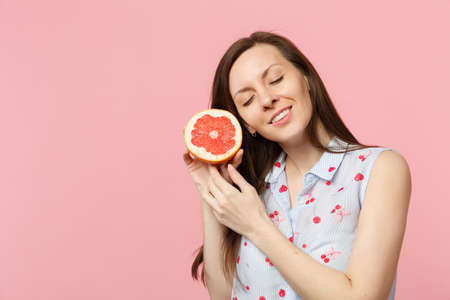 Pretty young woman in summer clothes keeping eyes closed, holding half of fresh ripe grapefruit isolated on pink pastel background. People vivid lifestyle relax vacation concept. Mock up copy spaceの写真素材