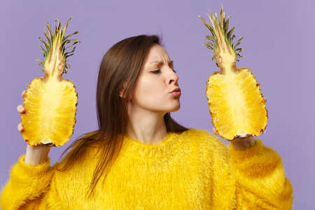 Cute young woman in fur sweater blowing sending air kiss hold halfs of fresh pineapple fruit isolated on violet pastel background. People vivid lifestyle, relax vacation concept. Mock up copy spaceの写真素材